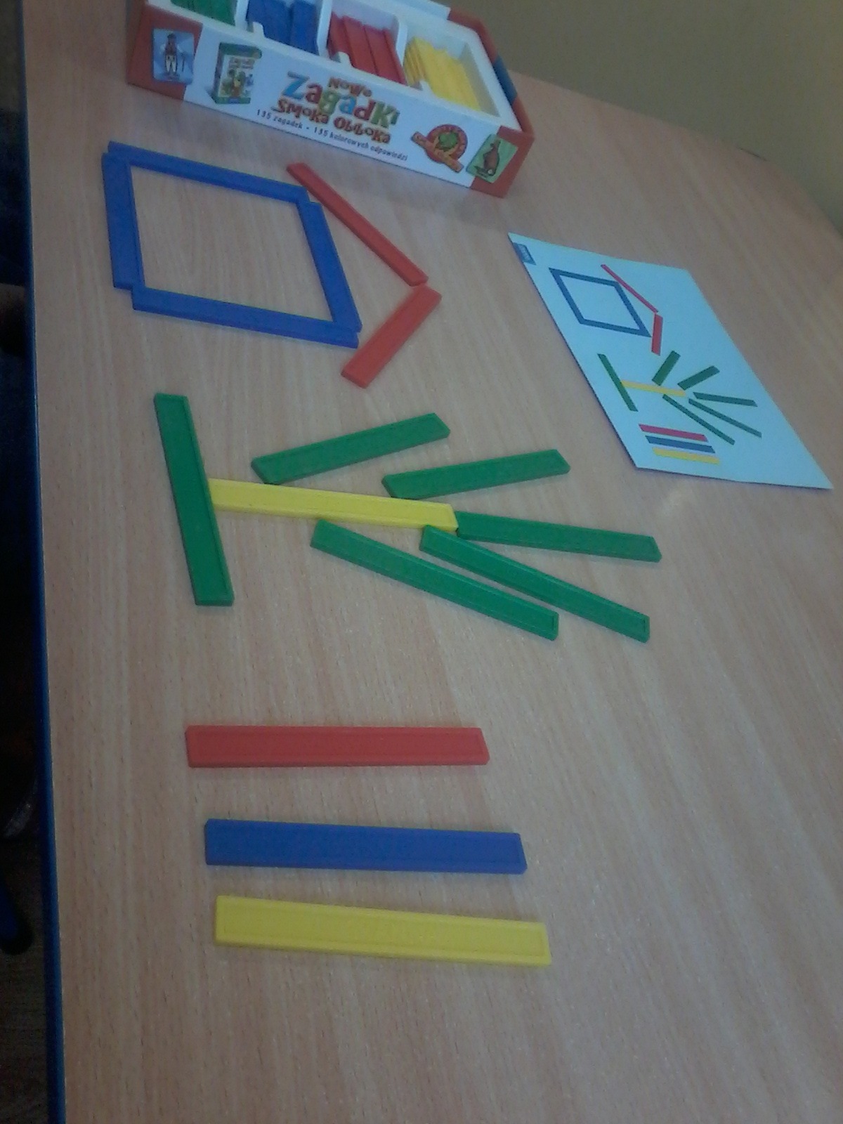 Rozsypane kolorowe klocki drewniane układane według wzoru z kartki, zestaw 'Nowe Zagadki Smoka Obiboka' na drewnianym stole.