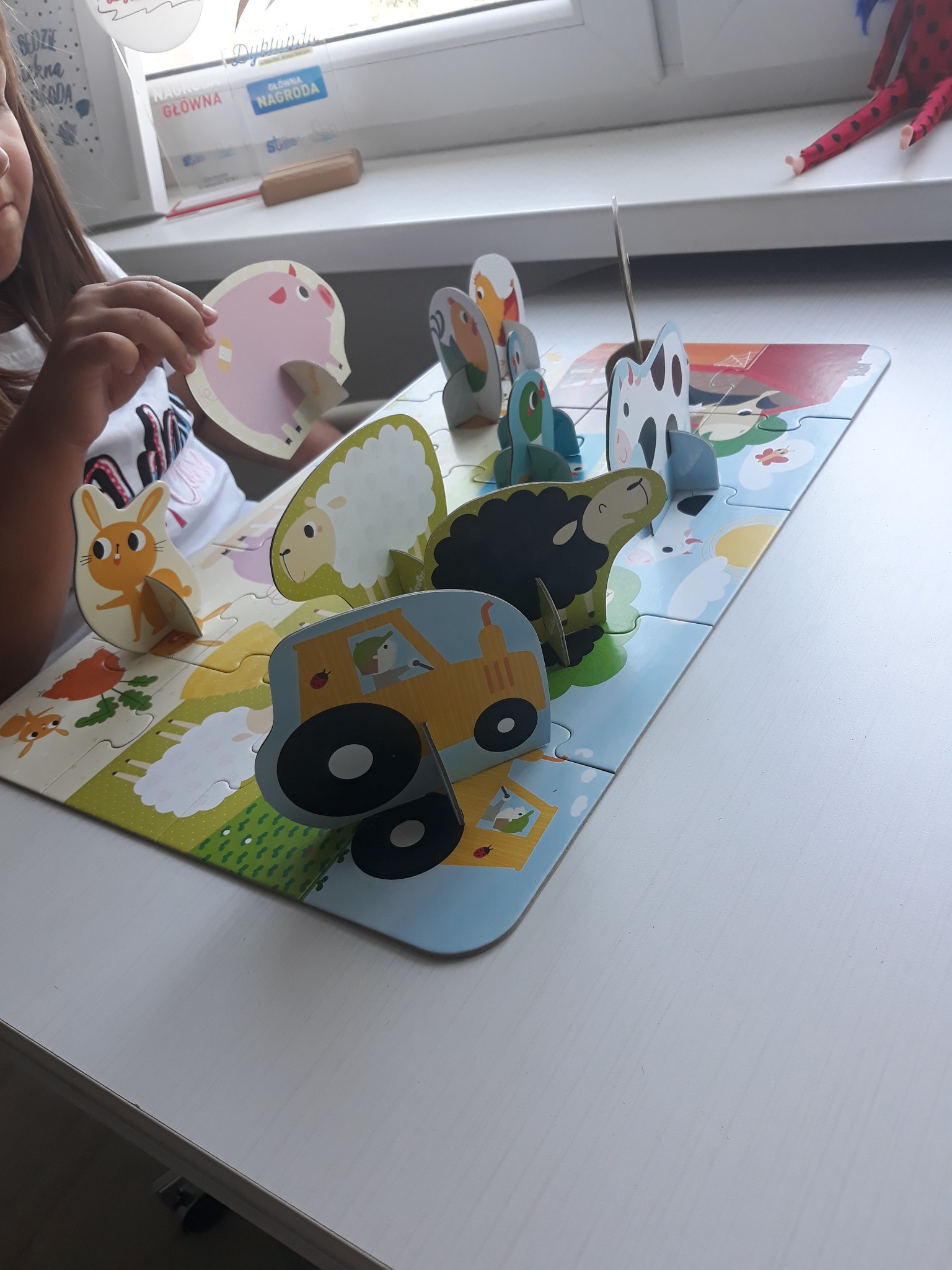 Dziewczynka układa trójwymiarowe puzzle z motywem farmy i zwierząt gospodarskich na białym stole przy oknie.