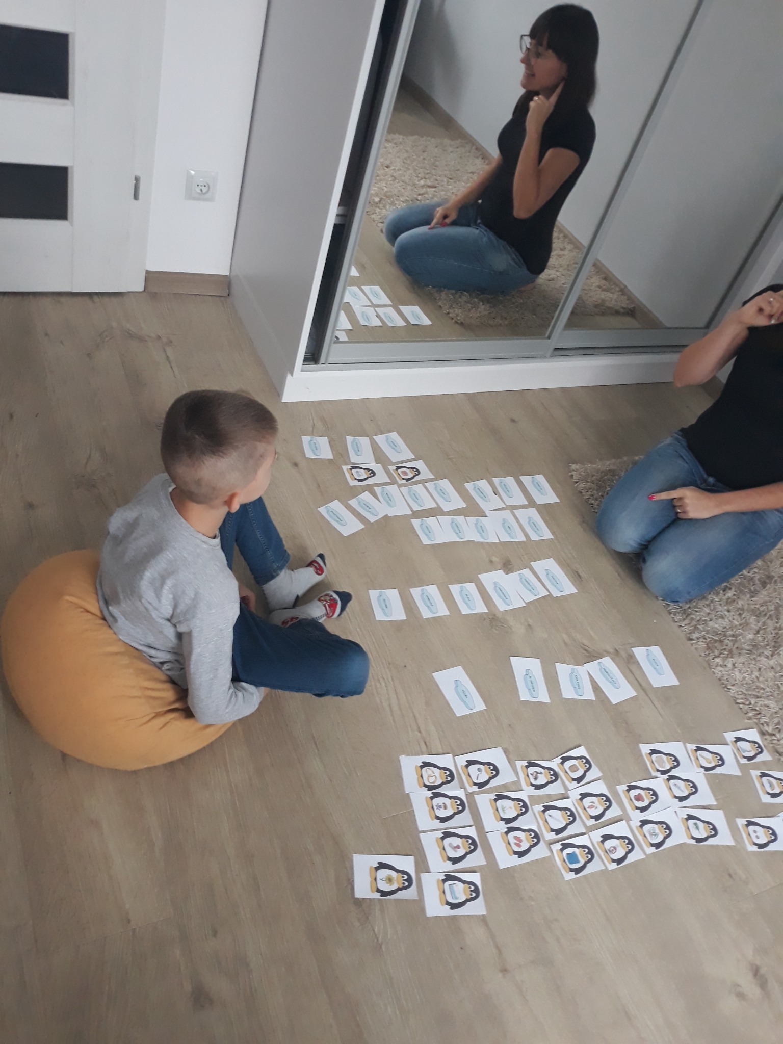 Chłopiec siedzi na żółtej pufie, gra w memory z kartami z pingwinami, kobieta w okularach siedzi na podłodze obok i wskazuje palcem na karty, odbicie w lustrze.
