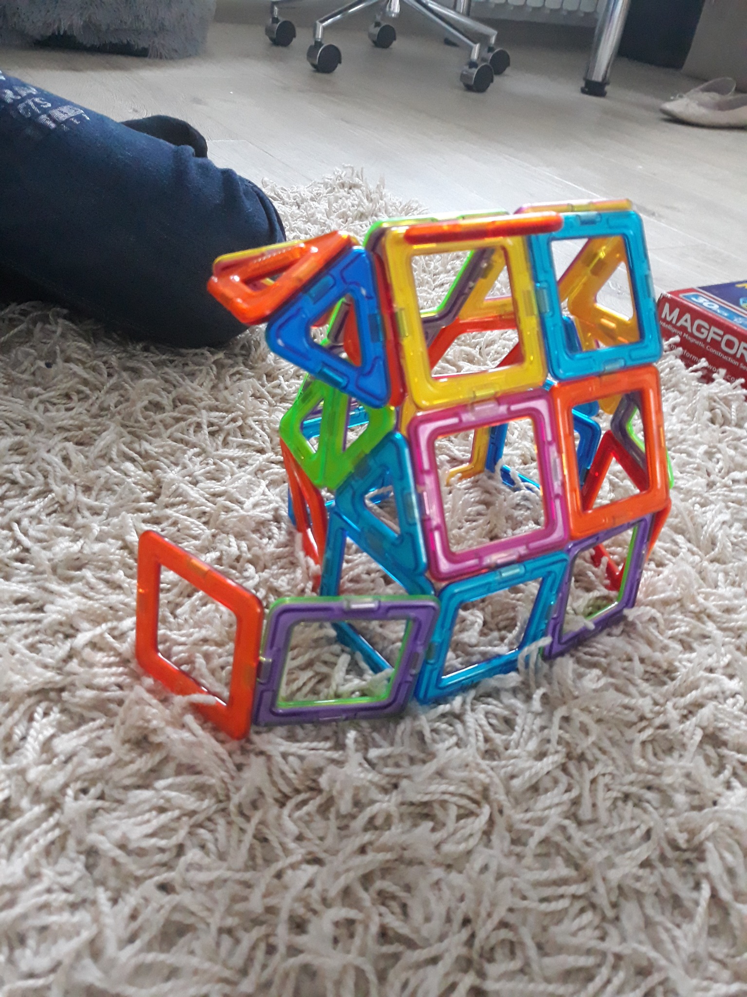 Kolorowa konstrukcja z magnetycznych klocków na puszystym dywanie, obok leżą niebieskie dżinsy i pudełko Magformers.