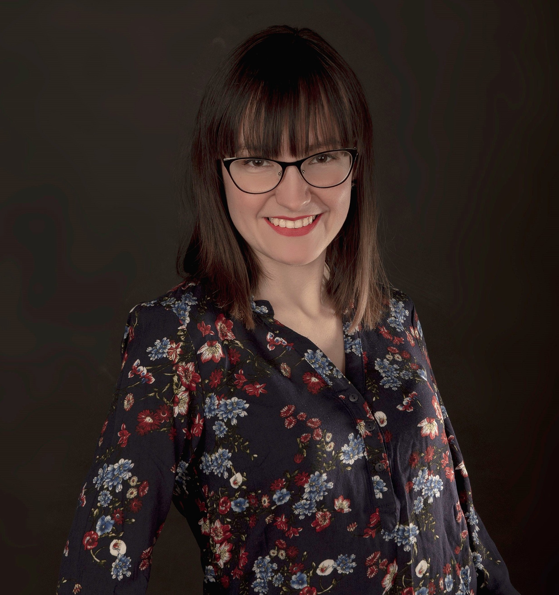 Uśmiechnięta kobieta w okularach i bluzce w kwiaty na ciemnym tle, prezentująca ciepły i profesjonalny wizerunek.