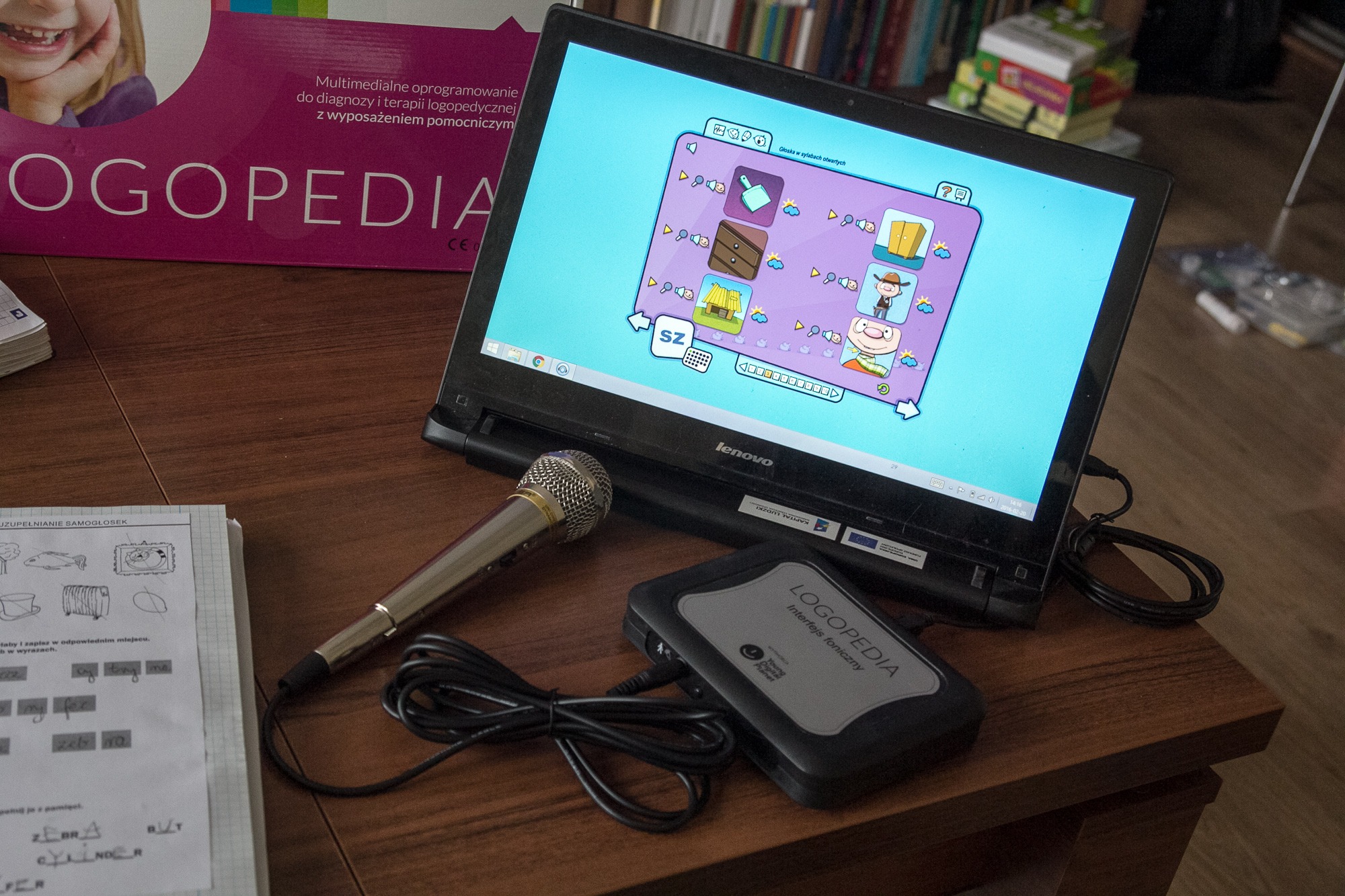 Stanowisko do terapii mowy z laptopem wyświetlającym interaktywną aplikację edukacyjną, mikrofonem i interfejsem fonicznym na drewnianym biurku, obok zeszyt z ćwiczeniami uzupełniania samogłosek.