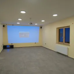 Pusta sala z szarymi płytkami podłogowymi, projektorem sufitowym wyświetlającym ekran testowy na ścianie, oświetleniem LED w suficie, grzejnikiem pod oknem i głośnikami na ścianach.