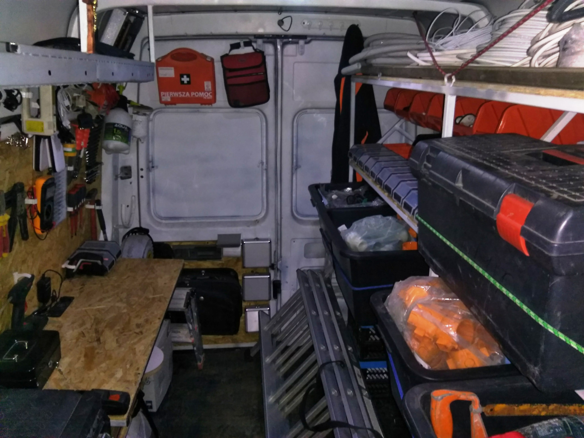 Wnętrze furgonetki serwisowej z półkami i narzędziami: skrzynki narzędziowe, drabina, akcesoria elektryczne, apteczka pierwszej pomocy, wiertarka na blacie roboczym z płyty OSB.