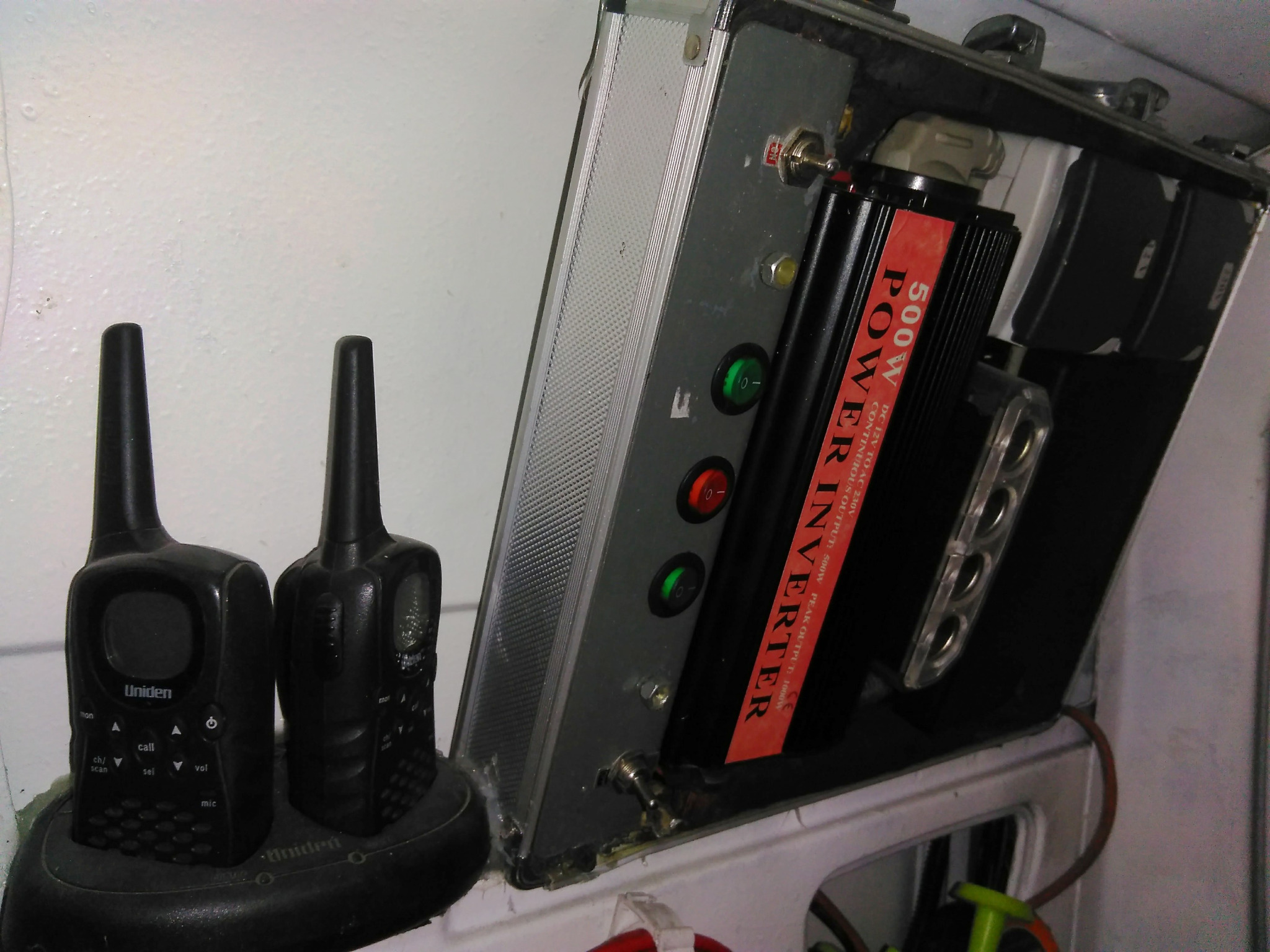 Dwa czarne radiotelefony Uniden obok srebrnej obudowy z zamontowanym czarnym inwerterem 500W z czerwoną etykietą, widoczne włączniki i gniazda.