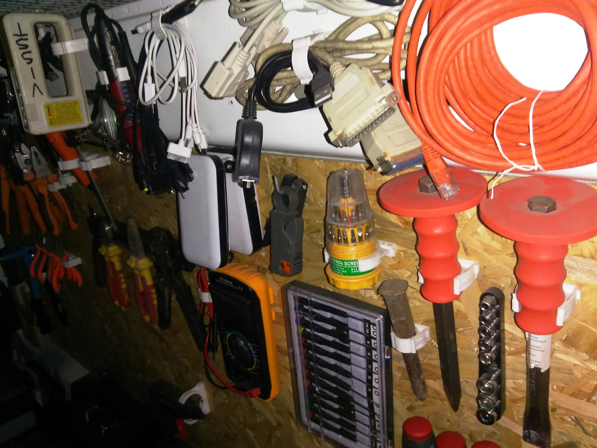 Ściana warsztatu z narzędziami elektrycznymi i pomiarowymi: multimetr, przewody, wkrętaki, dłuta, kombinerki, tester instalacji elektrycznej, wszystko przymocowane do ściany z płyty OSB za pomocą...
