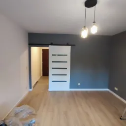 Nowoczesne wnętrze po remoncie: drewniana podłoga, szara ściana, białe drzwi przesuwne z poziomymi paskami, wiszące lampy. Widoczne farby malarskie.