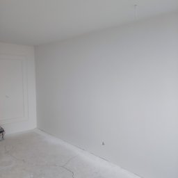 InwestAD Andrzej Dyki - Białe, świeżo wykończone wnętrze pokoju z widocznymi pęknięciami na podłodze i delikatną sztukaterią przy ścianie, przygotowane do dalszych prac wykończeniowych.