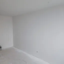 Białe, świeżo wykończone wnętrze pokoju z widocznymi pęknięciami na podłodze i delikatną sztukaterią przy ścianie, przygotowane do dalszych prac wykończeniowych.