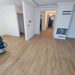 InwestAD Andrzej Dyki - Jasne panele podłogowe w nowym wnętrzu z kominkiem i gładkimi ścianami. Widoczny odkurzacz budowlany. W tle przejście do kolejnego pomieszczenia.