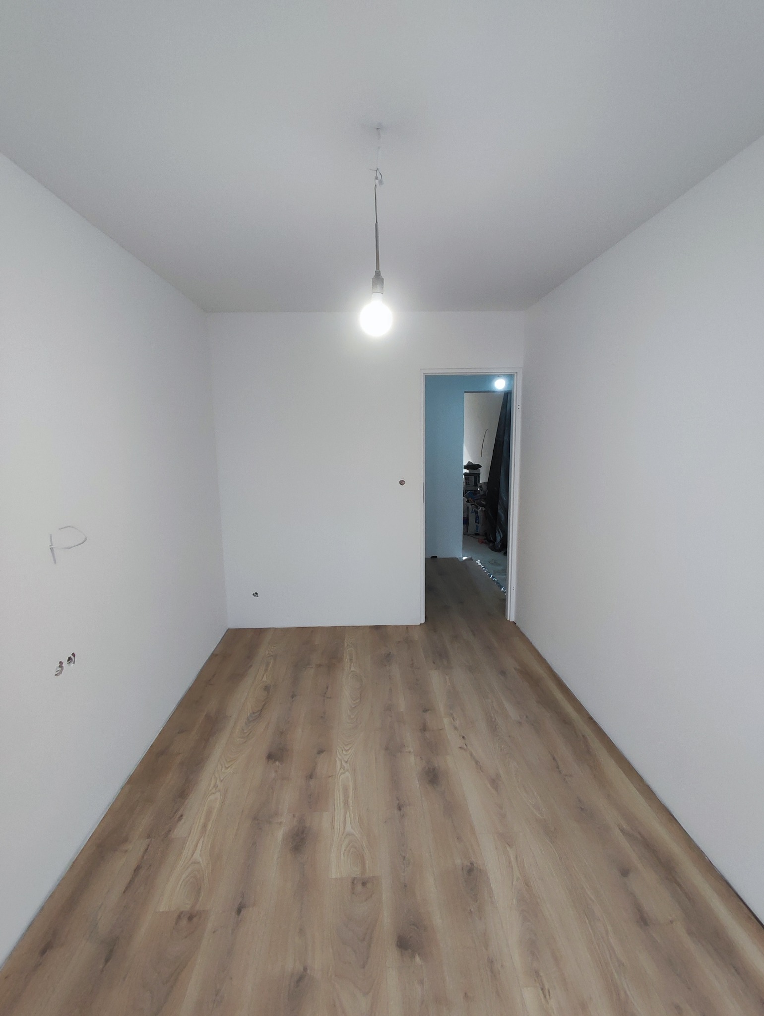 Wykończone wnętrze pokoju z drewnianą podłogą, świeżo pomalowanymi białymi ścianami i wiszącą żarówką. Widok na sąsiednie pomieszczenie.