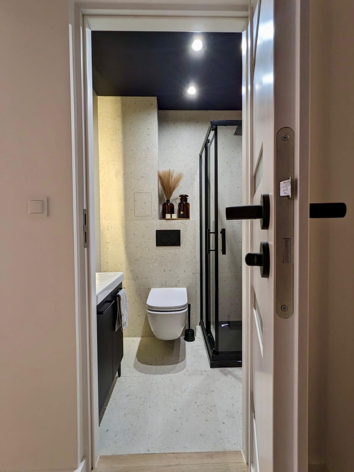 Nowoczesna łazienka z czarną kabiną prysznicową, umywalką i toaletą. Beżowe płytki, ciemny sufit. Widok zza uchylonych drzwi.