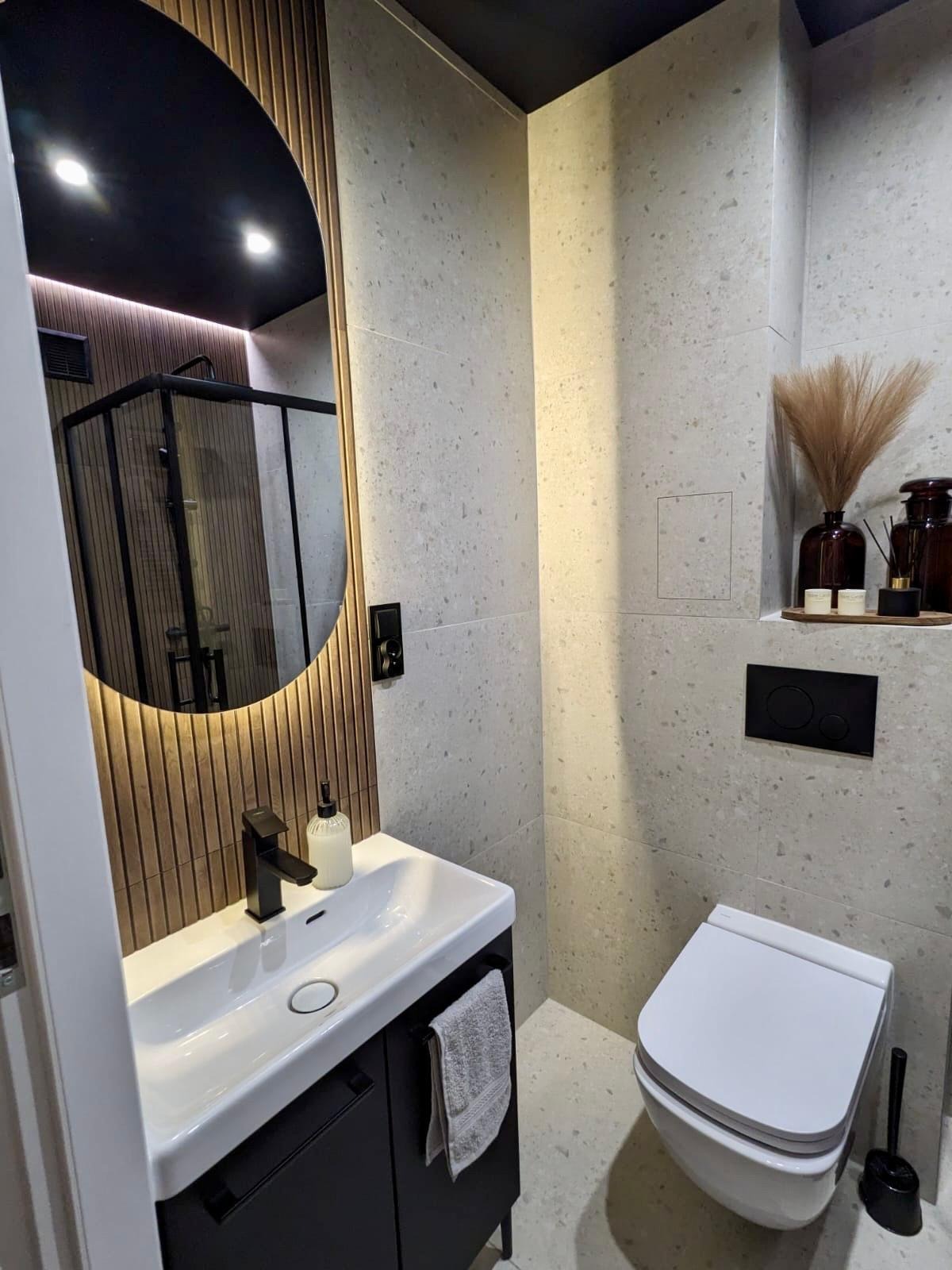 Nowoczesna łazienka z owalnym lustrem z podświetleniem LED, umywalką na szafce i toaletą. Płytki imitujące lastryko na ścianach i podłodze.