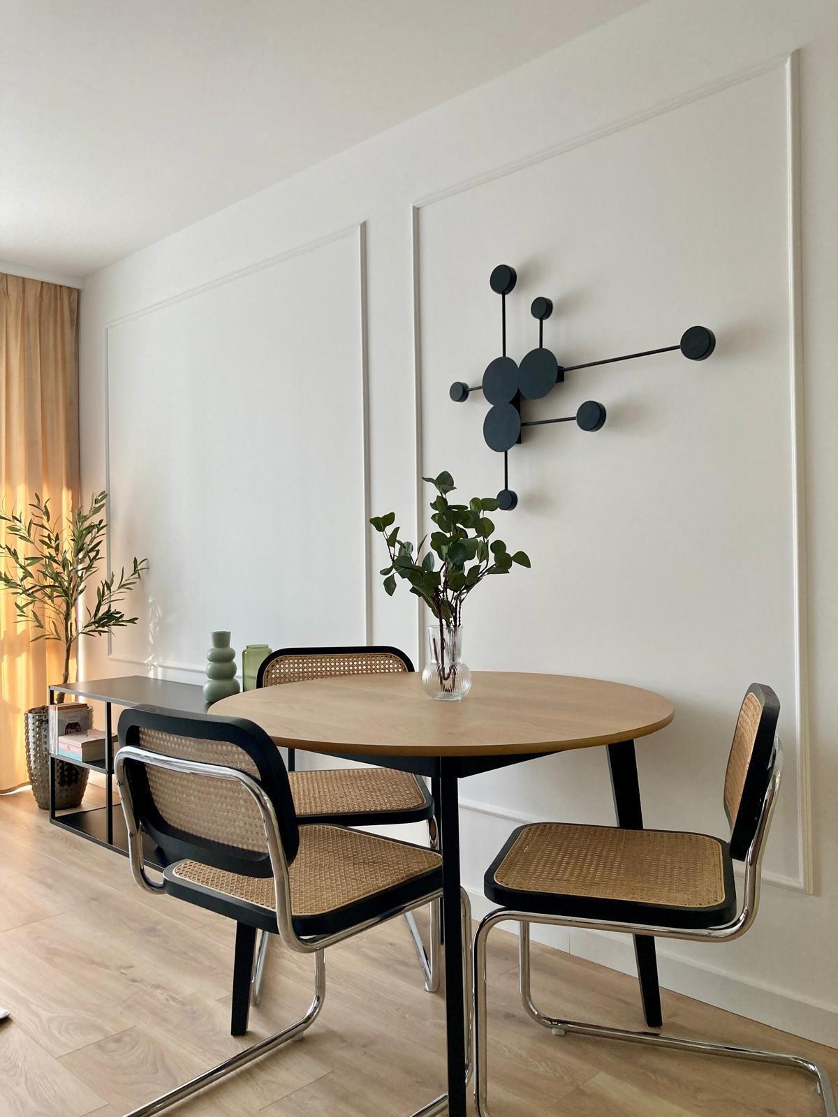 Stylowa jadalnia: okrągły stół z bukietem w wazonie, rattanowe krzesła z metalowymi nogami, designerski zegar na ścianie z dekoracyjnymi panelami.
