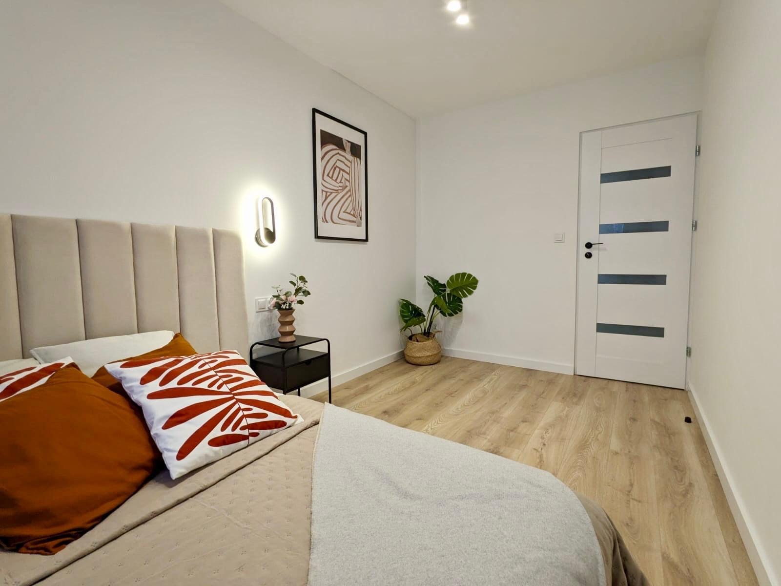 Jasna sypialnia z beżowym łóżkiem, wzorzystymi poduszkami i drewnianą podłogą. Na ścianie grafika w czarnej ramie i lampka. W rogu stoi roślina w plecionym koszu.