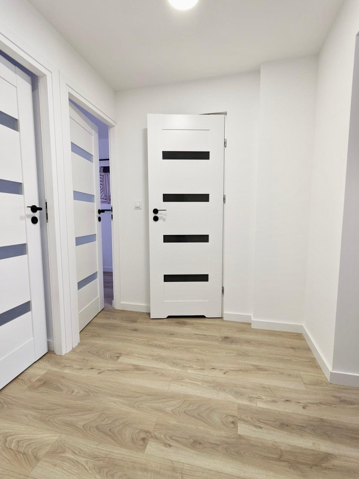 Jasne panele podłogowe w korytarzu z trzema białymi drzwiami z czarnymi akcentami. Jedne drzwi są otwarte, ukazując wnętrze pomieszczenia.