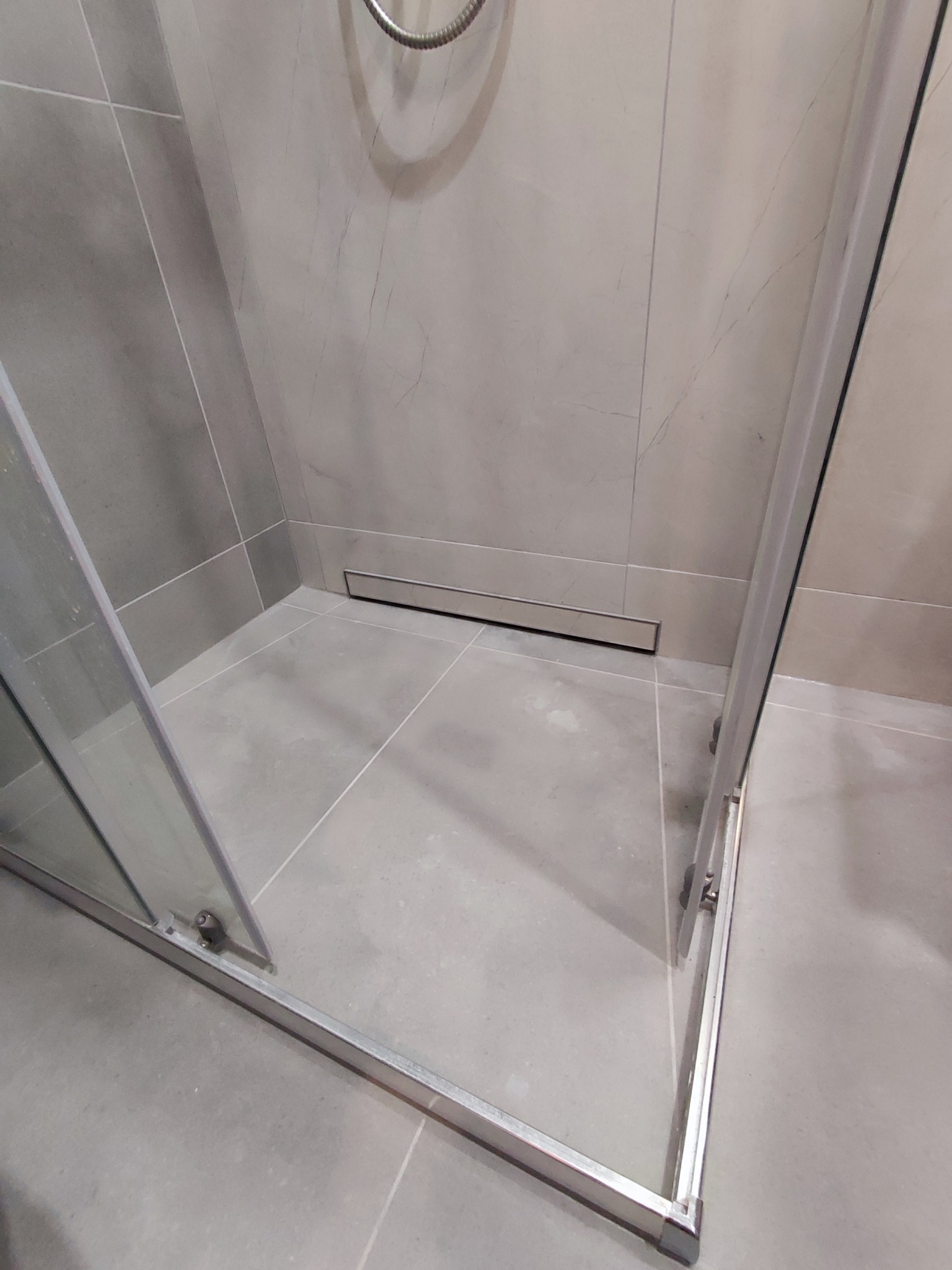 Szklana kabina prysznicowa z odpływem liniowym i szarymi płytkami imitującymi marmur, widziana z perspektywy wnętrza kabiny. Minimalistyczny design.