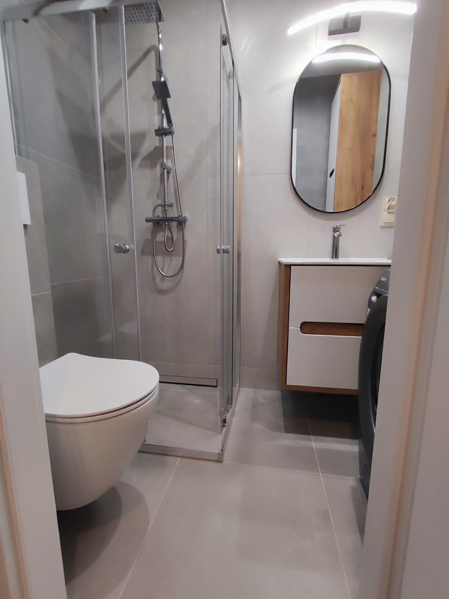 Nowoczesna łazienka w Tyszowcach po remoncie: kabina prysznicowa ze szklanymi drzwiami, wisząca toaleta, umywalka z szafką i owalne lustro z oświetleniem LED.