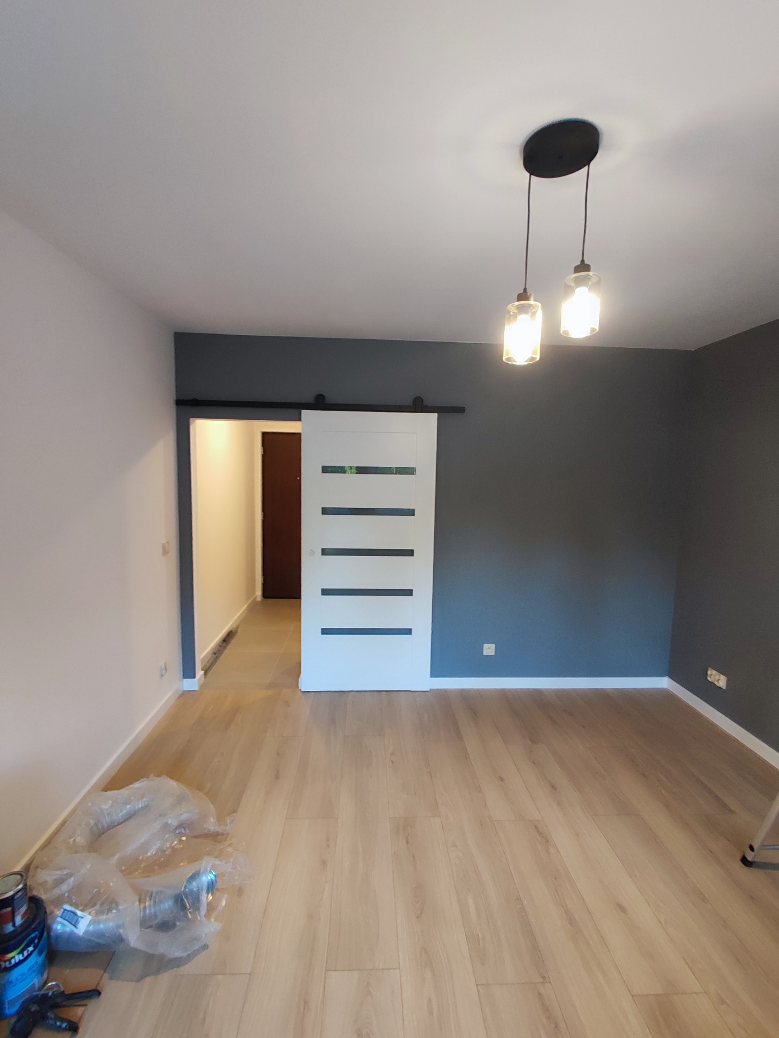 Nowoczesne wnętrze po remoncie: drewniana podłoga, szara ściana, białe drzwi przesuwne z poziomymi paskami, wiszące lampy. Widoczne farby malarskie.