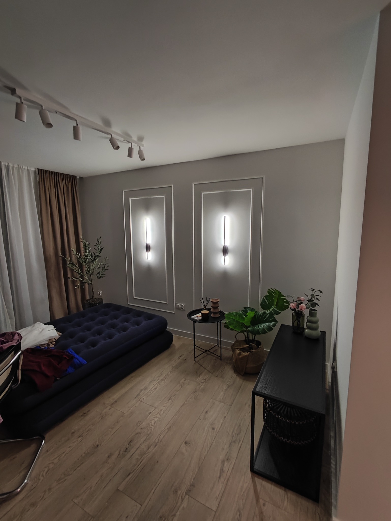 Nowoczesne wnętrze: drewniana podłoga, designerskie lampy ścienne z ramami, ciemny materac, rośliny doniczkowe i minimalistyczne meble. Oświetlenie punktowe na suficie.