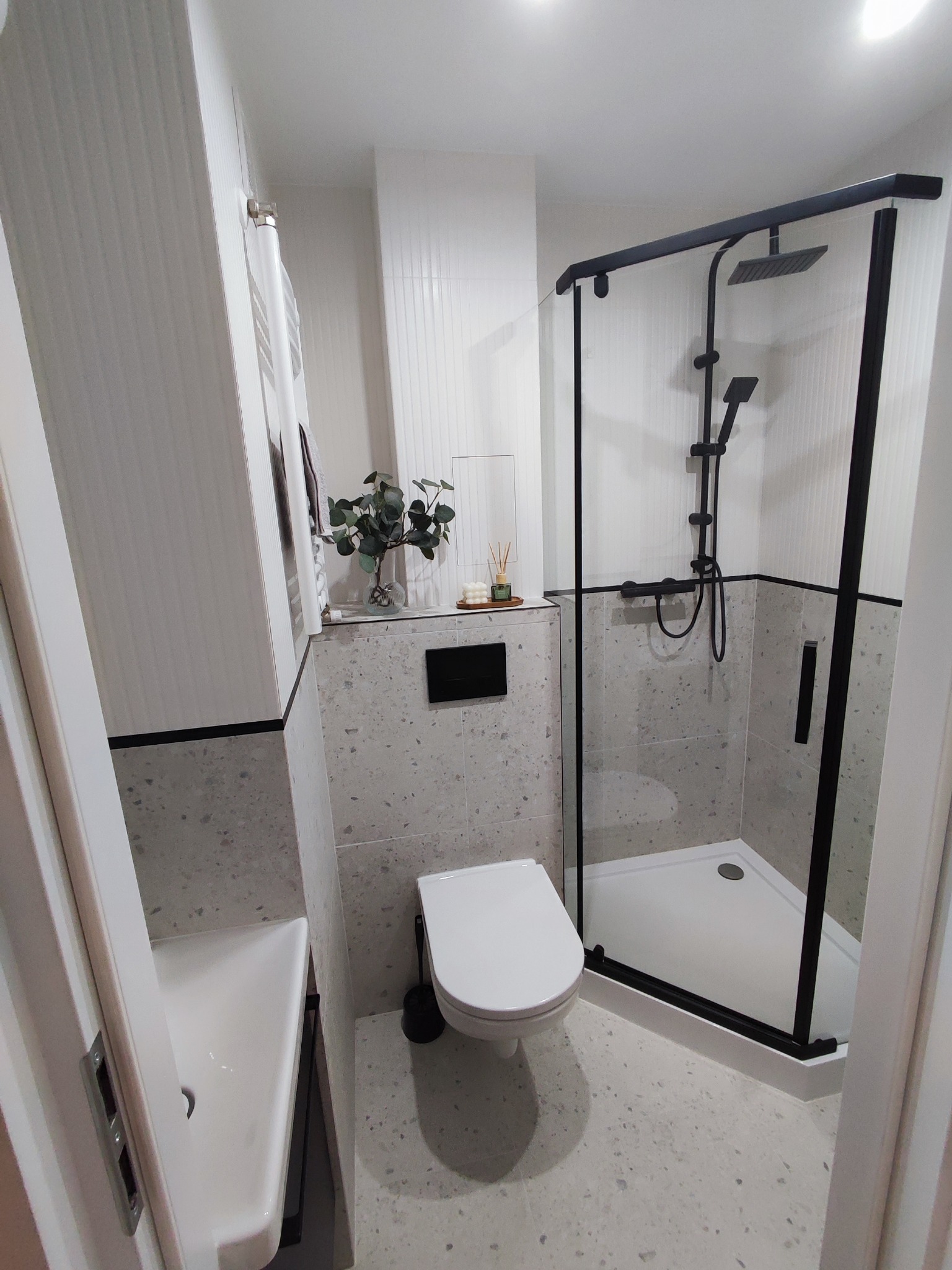 Nowoczesna łazienka z kabiną prysznicową w czarnej ramie, umywalką i toaletą. Ściany i podłoga wykończone płytkami imitującymi lastryko. Minimalistyczny design.