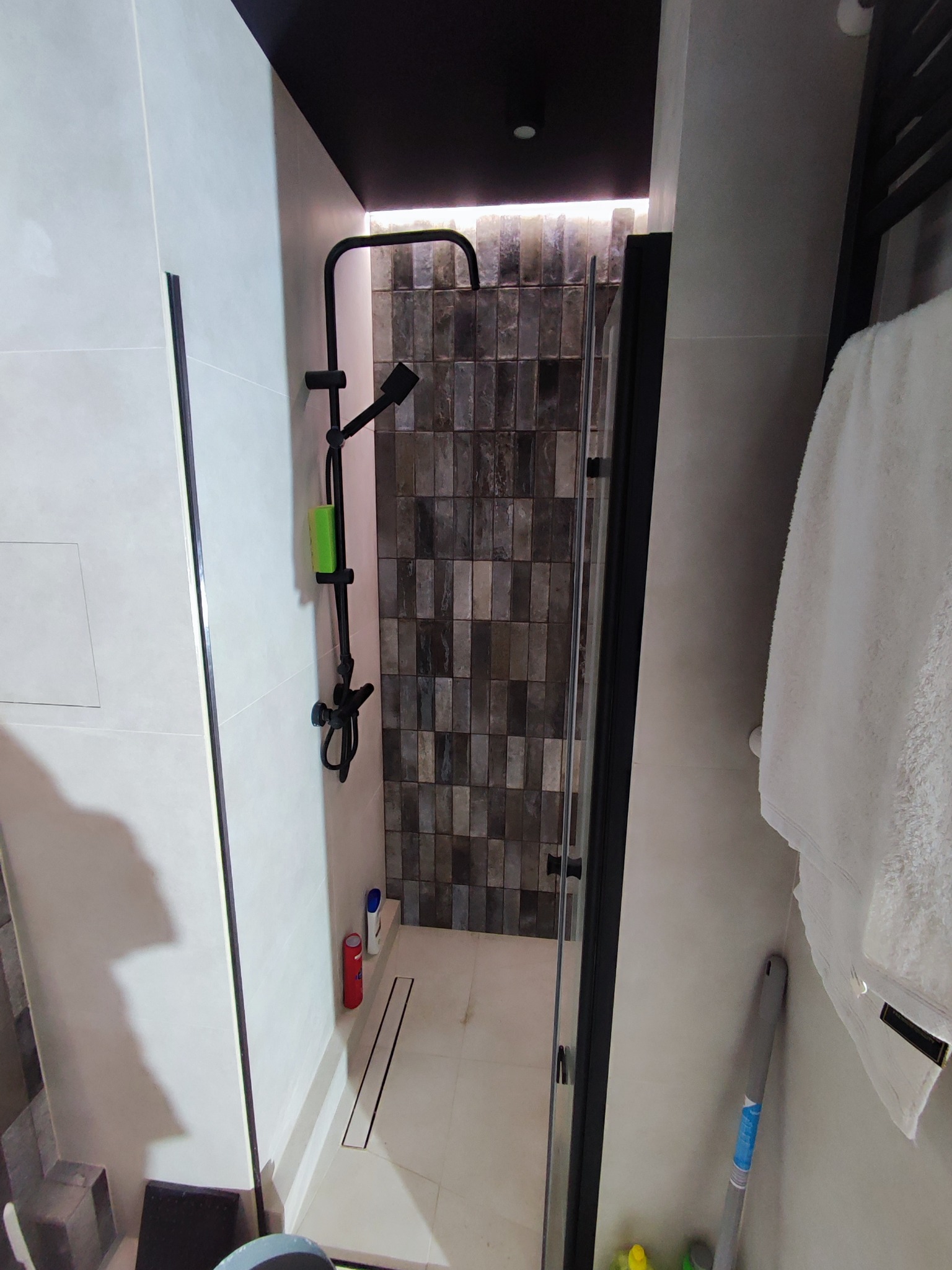 Nowoczesna kabina prysznicowa z czarną armaturą i odpływem liniowym. Ściany wyłożone płytkami w odcieniach szarości. Biały ręcznik na grzejniku.