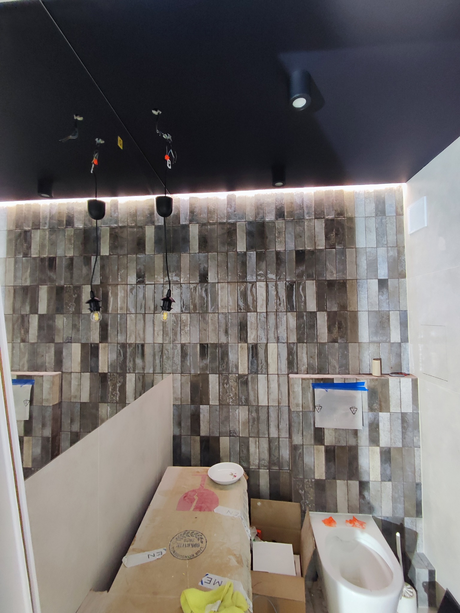 Wykończona ściana z szarych, podłużnych płytek w łazience. Widoczne wiszące żarówki bez kloszy, czarny sufit i częściowo zakryta toaleta.