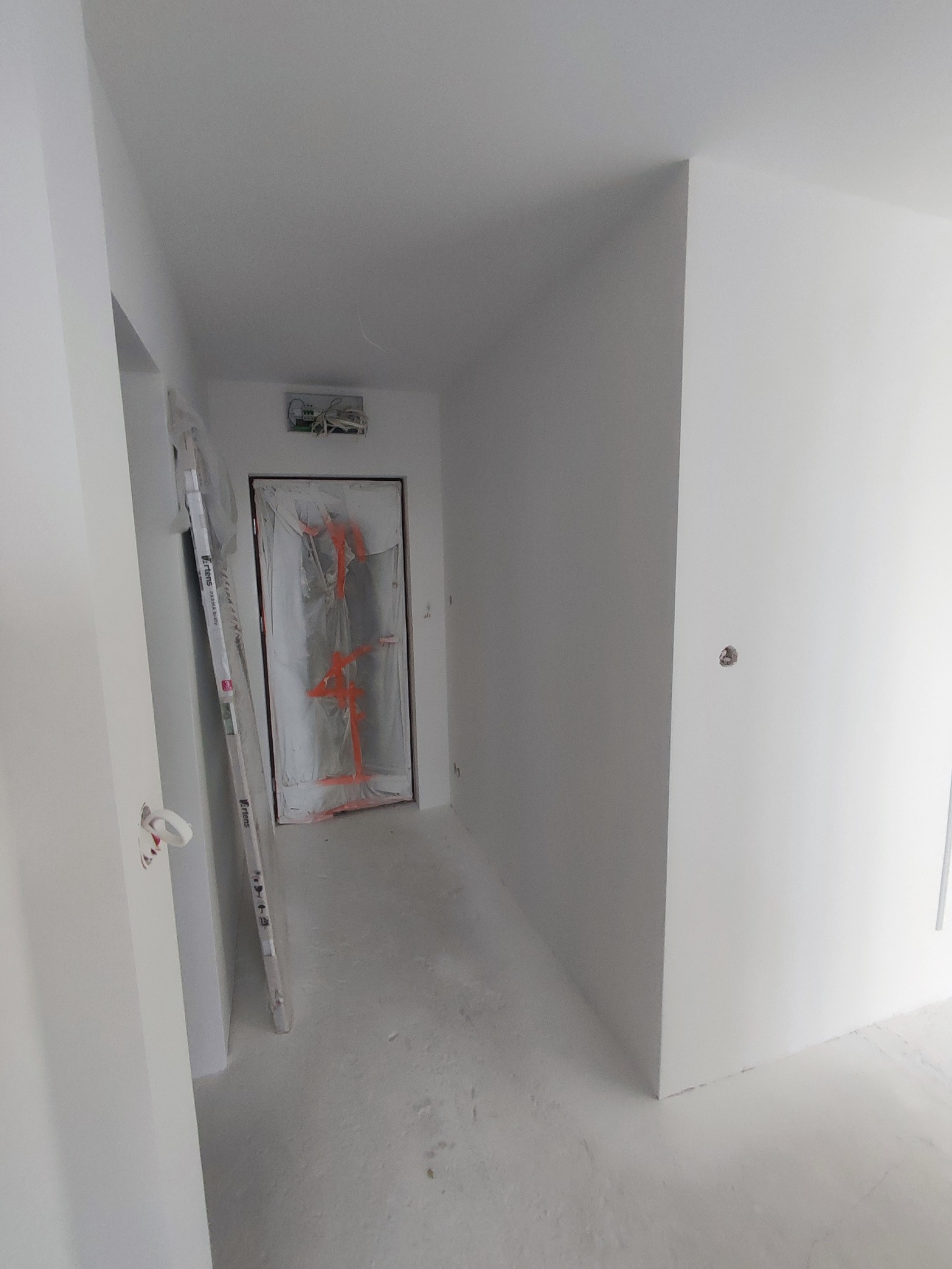Białe, świeżo pomalowane wnętrze korytarza z zabezpieczonymi drzwiami folią malarską. Widoczne listwy przypodłogowe i ślady farby na podłodze.