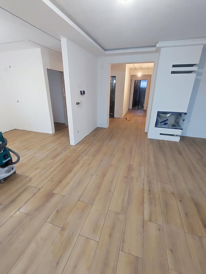 Jasne panele podłogowe w nowym wnętrzu z kominkiem i gładkimi ścianami. Widoczny odkurzacz budowlany. W tle przejście do kolejnego pomieszczenia.