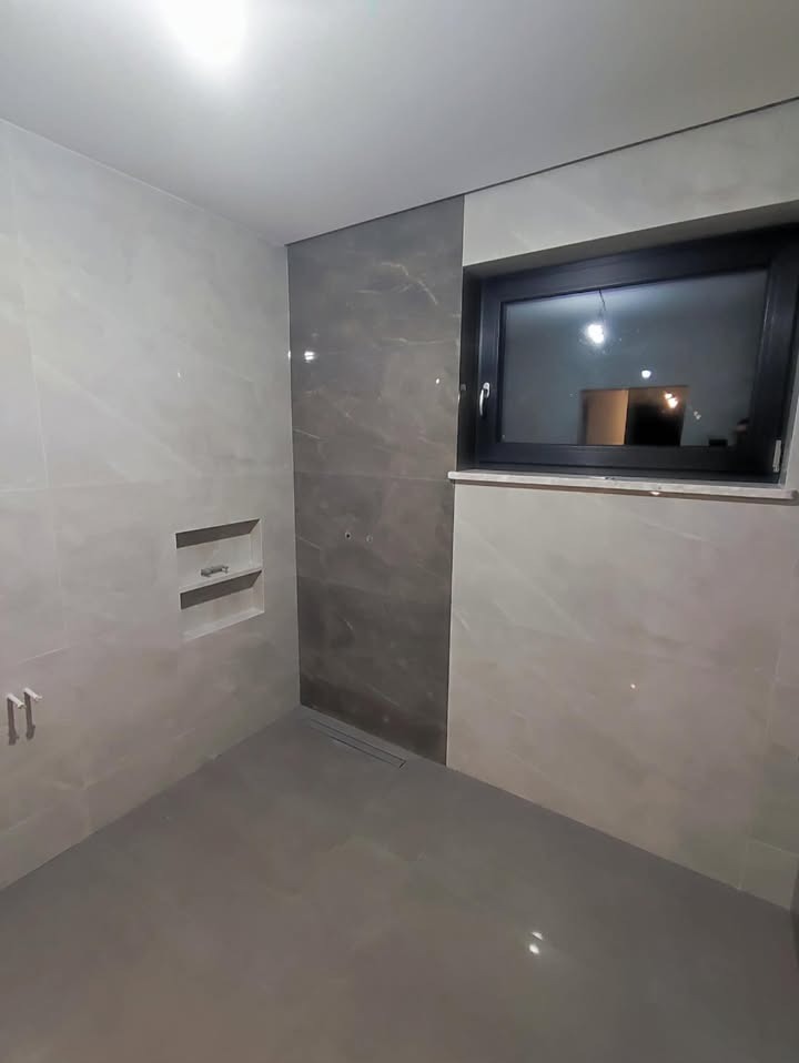 Wykończona łazienka z szarymi płytkami imitującymi marmur, czarną ramą okna i wnęką na kosmetyki. Minimalistyczny design z odpływem liniowym.