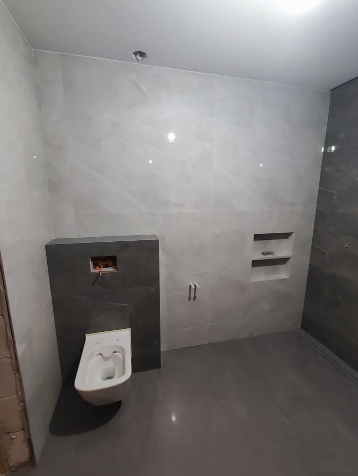 Nowoczesna łazienka w szarościach z podtynkową toaletą i wnęką w ścianie. Widoczne płytki wielkoformatowe i odpływ liniowy. Minimalistyczny design.