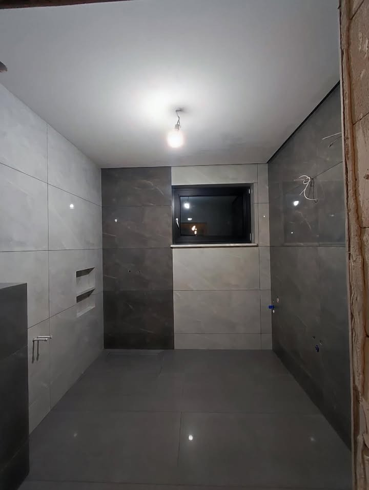 Nowoczesna łazienka z szarymi płytkami imitującymi marmur, czarnym oknem i minimalistycznym oświetleniem. Widoczne wnęki w ścianie i fragment surowej ściany.