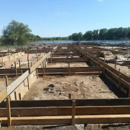 Szeroki kadr na przygotowanie terenu pod fundamenty z drewnianym szalunkiem, widok na rzekę i drzewa w tle pod błękitnym niebem.