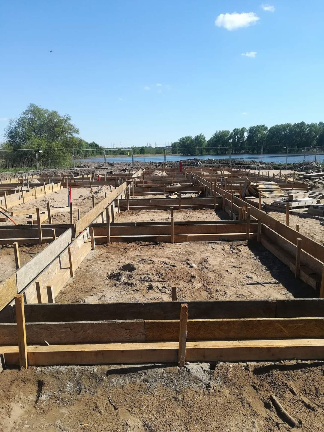 Szeroki kadr na przygotowanie terenu pod fundamenty z drewnianym szalunkiem, widok na rzekę i drzewa w tle pod błękitnym niebem.