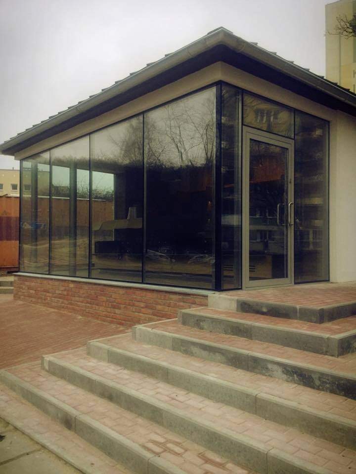 Szklana oranżeria z ceglaną podmurówką i schodami z kostki brukowej prowadzącymi do wejścia, widok zewnętrzny w pochmurny dzień.