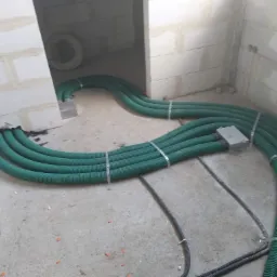 Wnętrze w stanie surowym z widocznymi zielonymi rurami wentylacyjnymi ułożonymi na podłodze i przymocowanymi metalowymi obejmami, w tle białe ściany z bloczków.