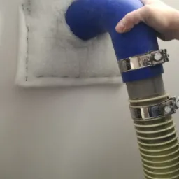 Wdmuchiwanie białej izolacji do poddasza za pomocą niebieskiego węża z metalowymi zaciskami, widoczna faktura materiału izolacyjnego i szew na krawędzi.