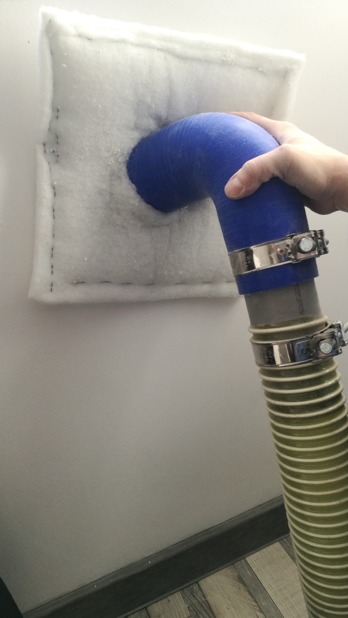 Wdmuchiwanie białej izolacji do poddasza za pomocą niebieskiego węża z metalowymi zaciskami, widoczna faktura materiału izolacyjnego i szew na krawędzi.