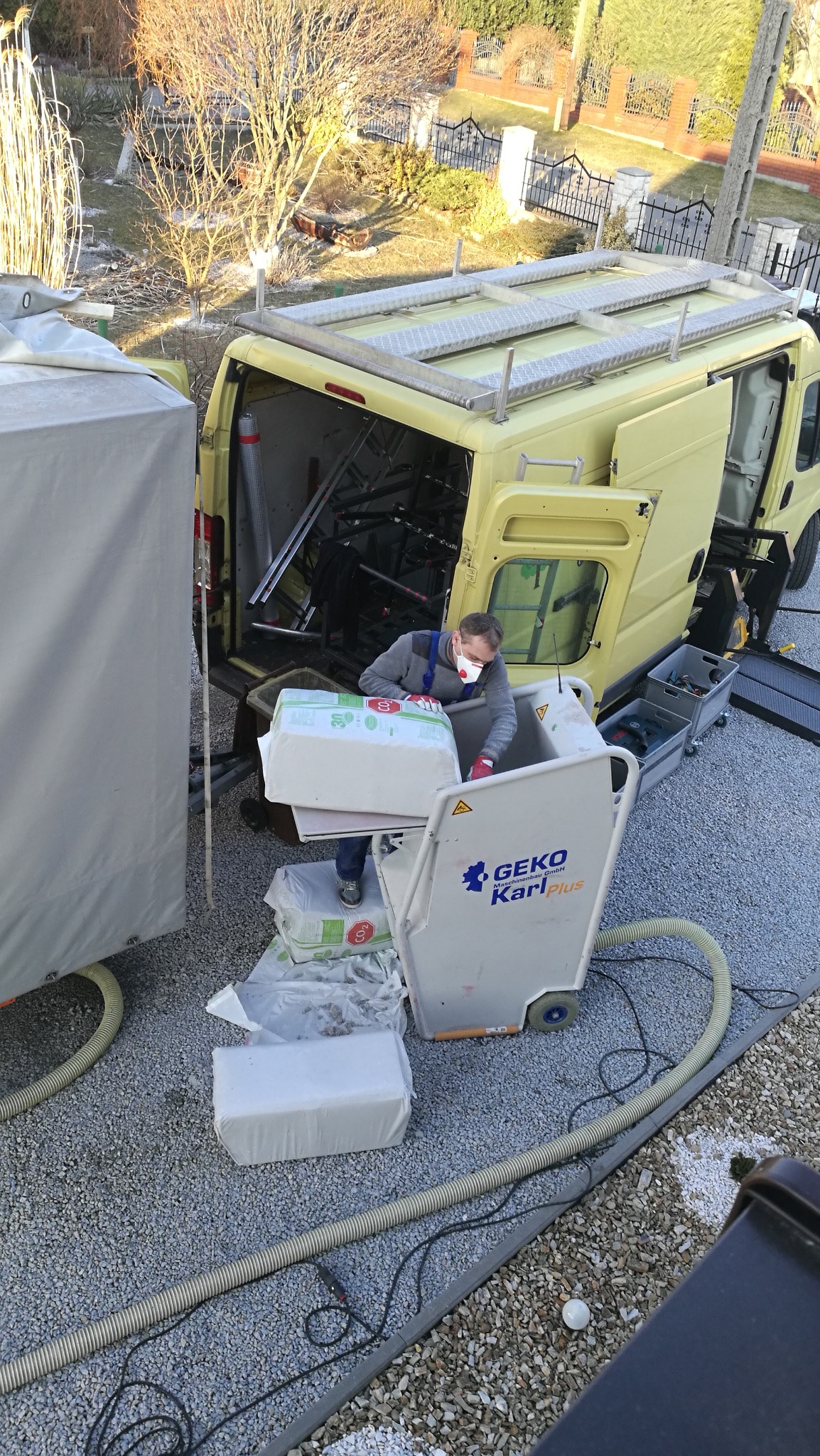 Pracownik w masce i rękawicach przenosi pakiet izolacji z żółtego samochodu dostawczego do maszyny Geko Karl Plus na żwirowym podłożu.