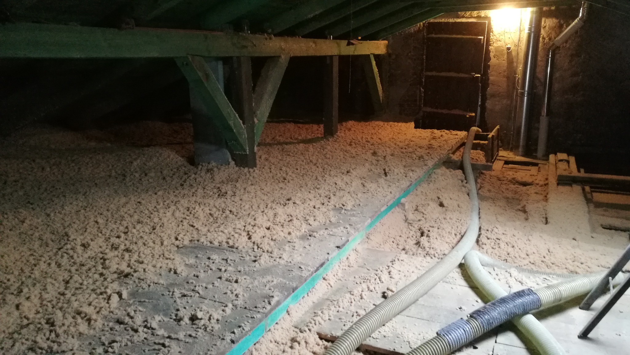 Poddasze z widoczną warstwą izolacji w postaci wełny celulozowej, zielony element konstrukcyjny oraz rury wentylacyjne w tle.