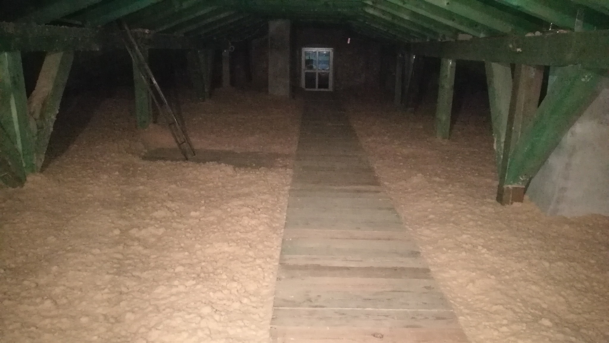 Poddasze z widoczną izolacją z wełny mineralnej, drewnianą kładką i oknem w tle, oświetlone słabym światłem.