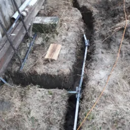 Wykop pod fundament z widoczną metalową konstrukcją wzmacniającą i pracownikiem wyrównującym dno, z widoczną deską i sznurkiem wyznaczającym linię.