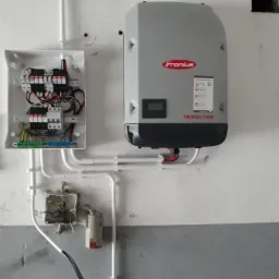 Szara obudowa falownika Fronius Symo oraz rozdzielnia elektryczna z zabezpieczeniami, zamontowane na pomalowanej ścianie w Biłgoraju, z widocznymi białymi rurami instalacyjnymi.