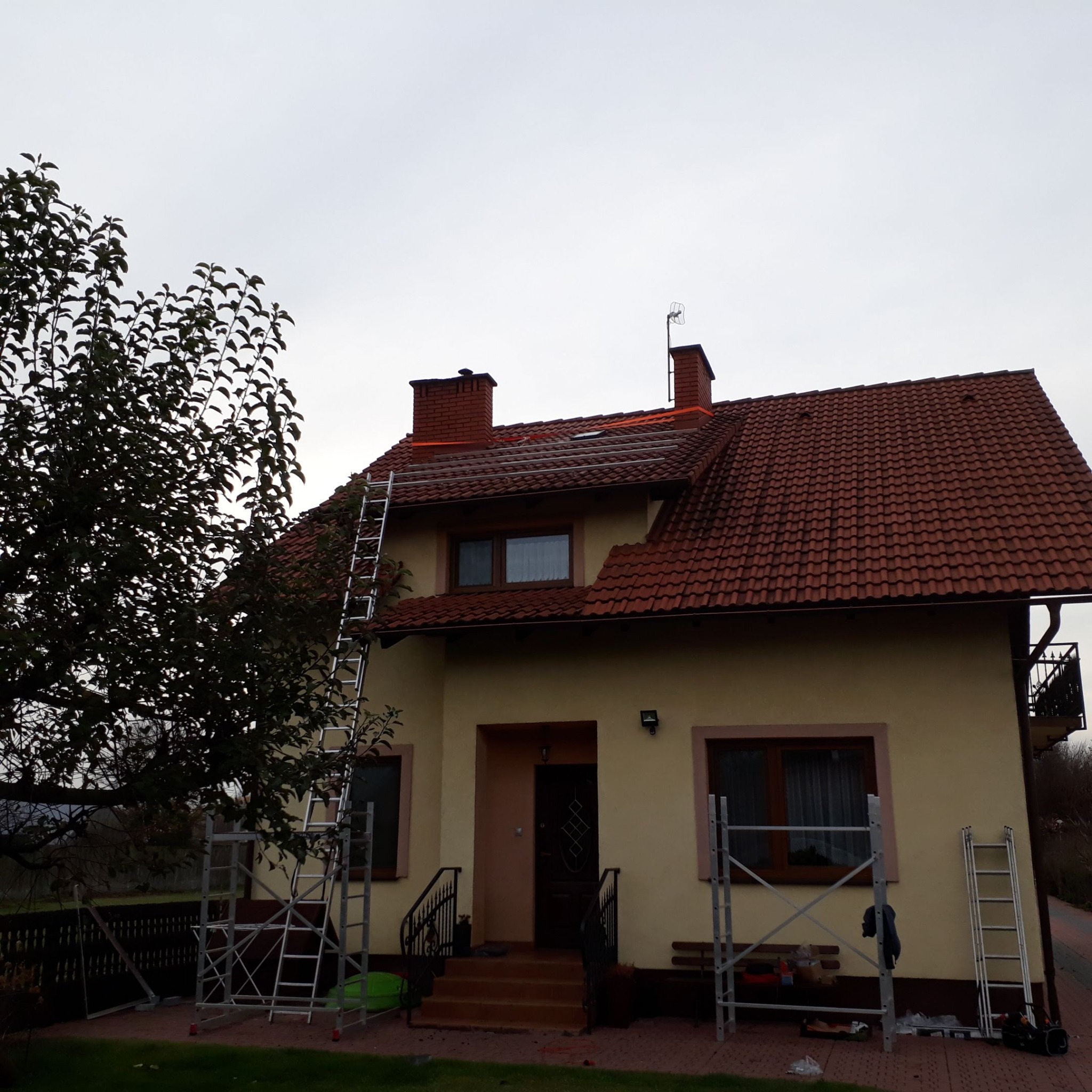 Dom jednorodzinny w Biłgoraju z czerwoną dachówką i rozstawionymi rusztowaniami oraz drabinami sugerującymi prace na wysokości, prawdopodobnie związane z dachem lub instalacjami na dachu.