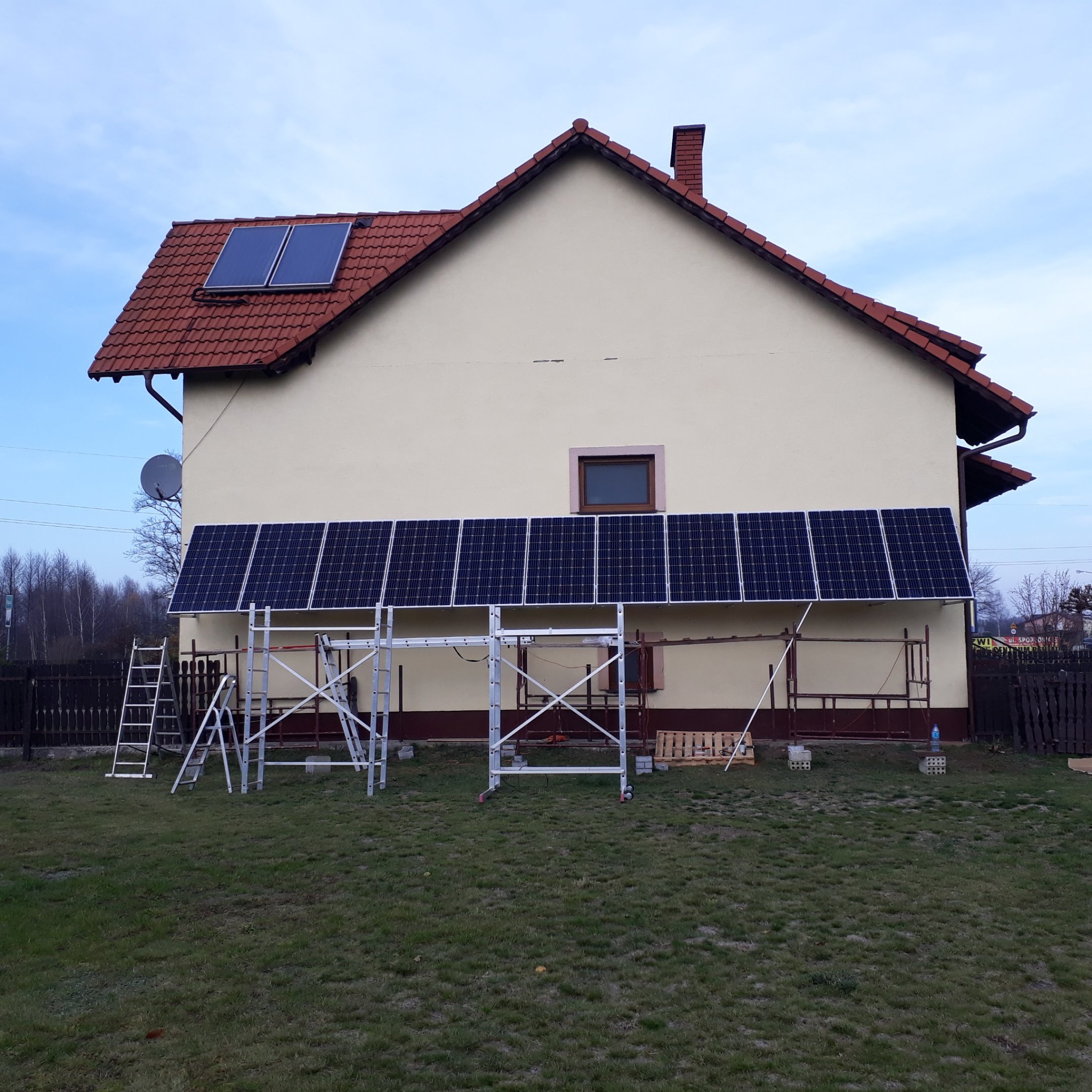 Montaż paneli słonecznych na ścianie domu jednorodzinnego, widoczne rusztowania i drabina, dwa panele na dachu.