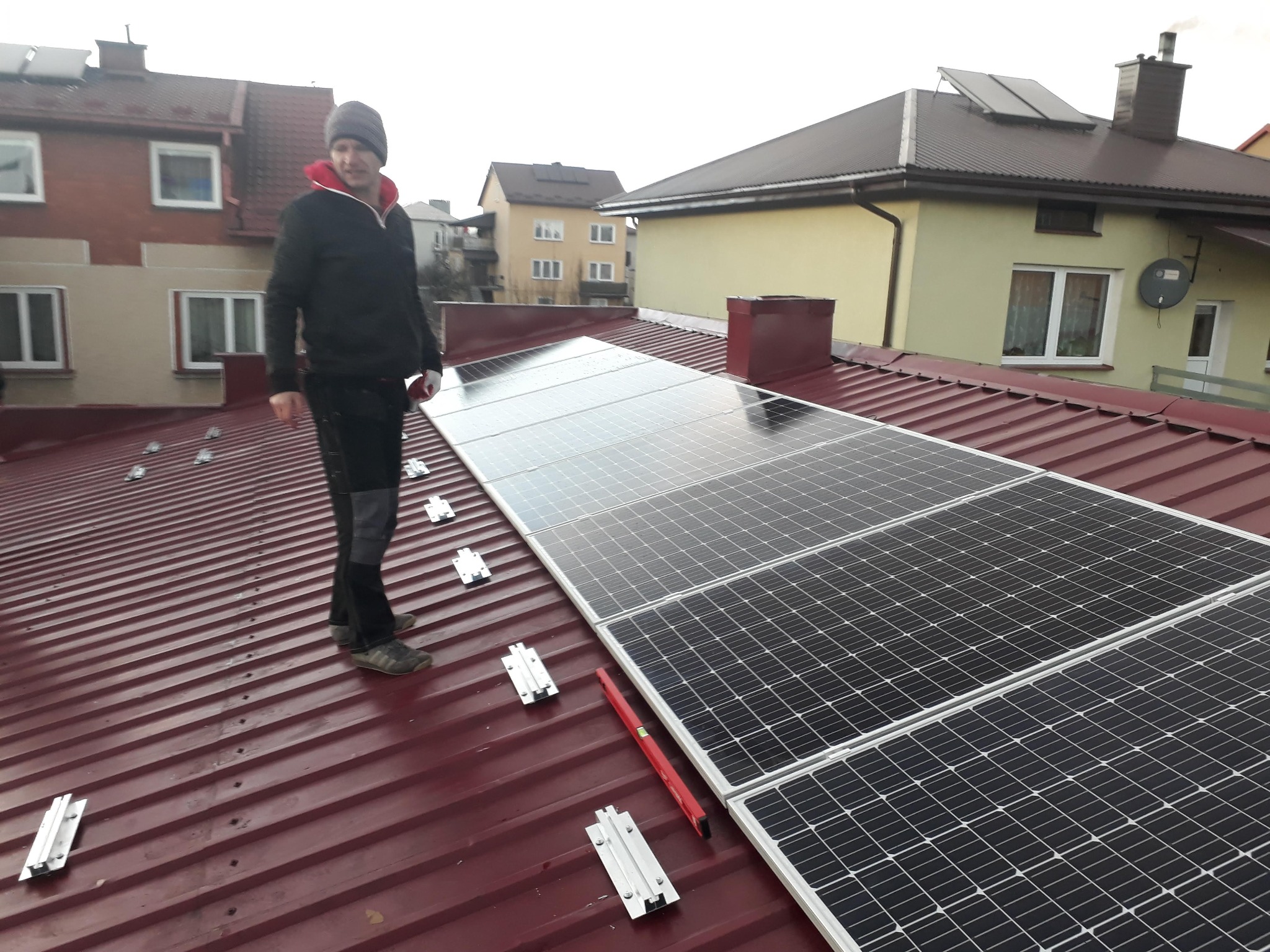 Montaż paneli słonecznych na dachu z blachy trapezowej; widoczny instalator w czapce i poziomica; w tle zabudowa jednorodzinna.