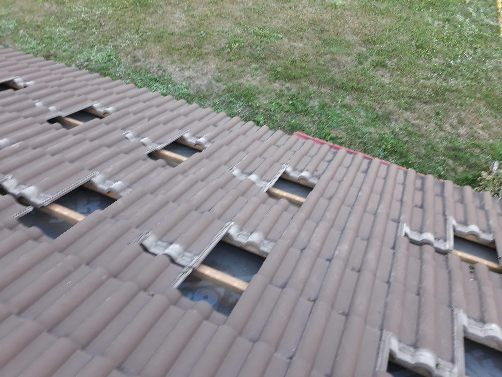 Fragment brązowego dachu z ceramiczną dachówką, miejscami zdemontowaną, odsłaniającą drewnianą konstrukcję, na tle zieleni.