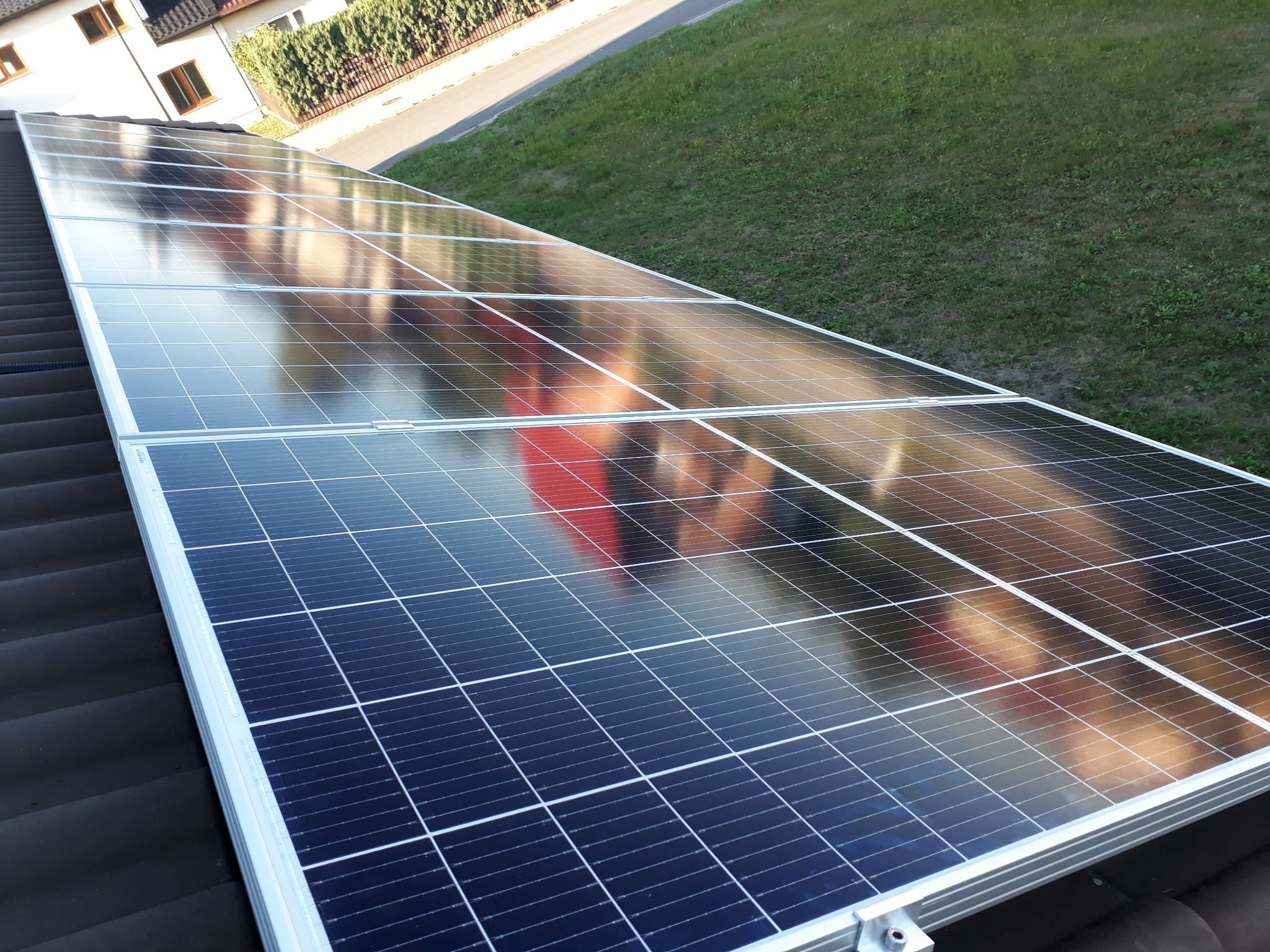 Panele słoneczne na brązowym dachu, odbijające otoczenie, z widocznym trawnikiem i fragmentem budynku w tle.