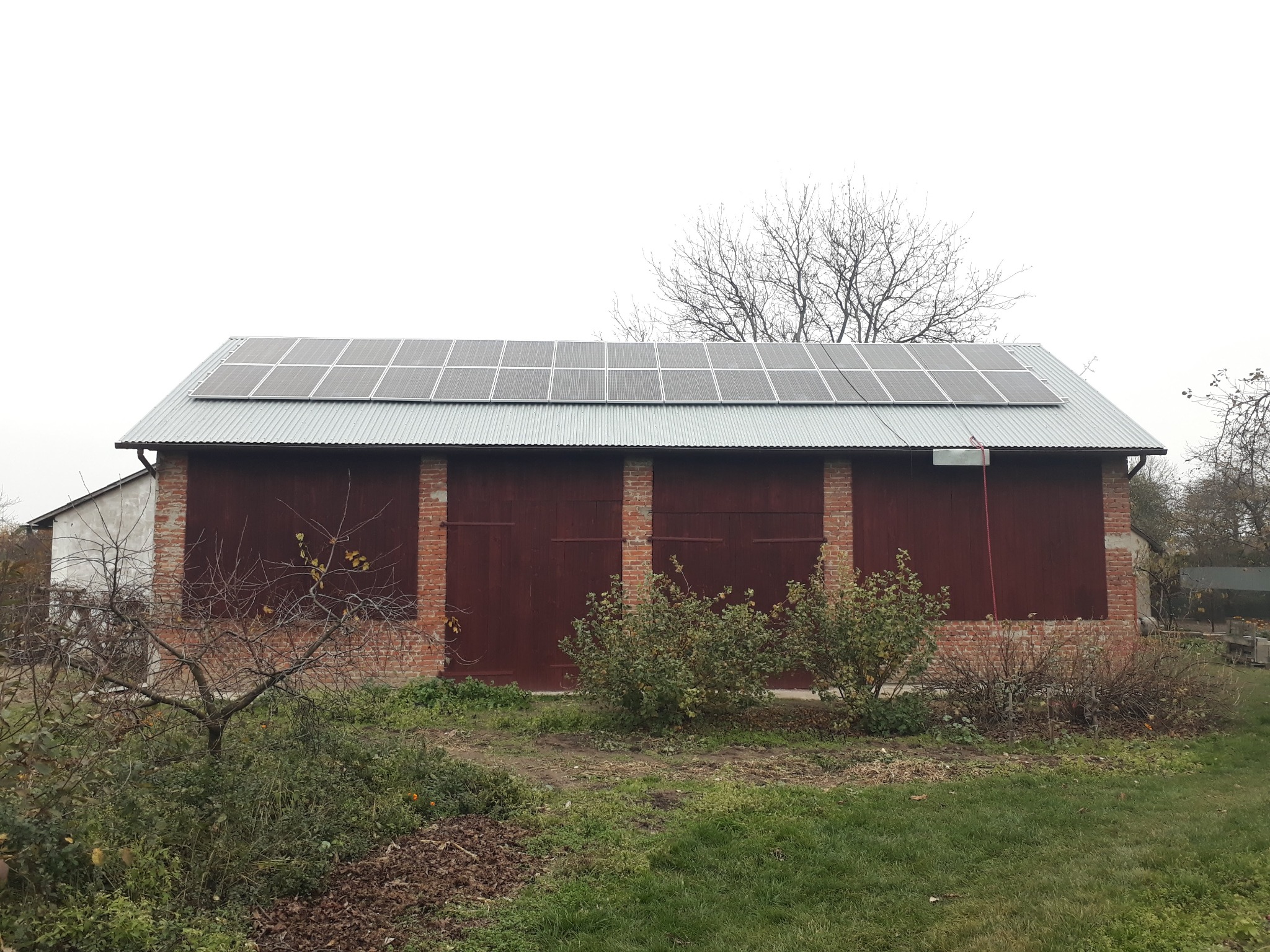Panele fotowoltaiczne zamontowane na blaszanym dachu budynku gospodarczego z czerwonej cegły, widok z poziomu gruntu w pochmurny dzień.