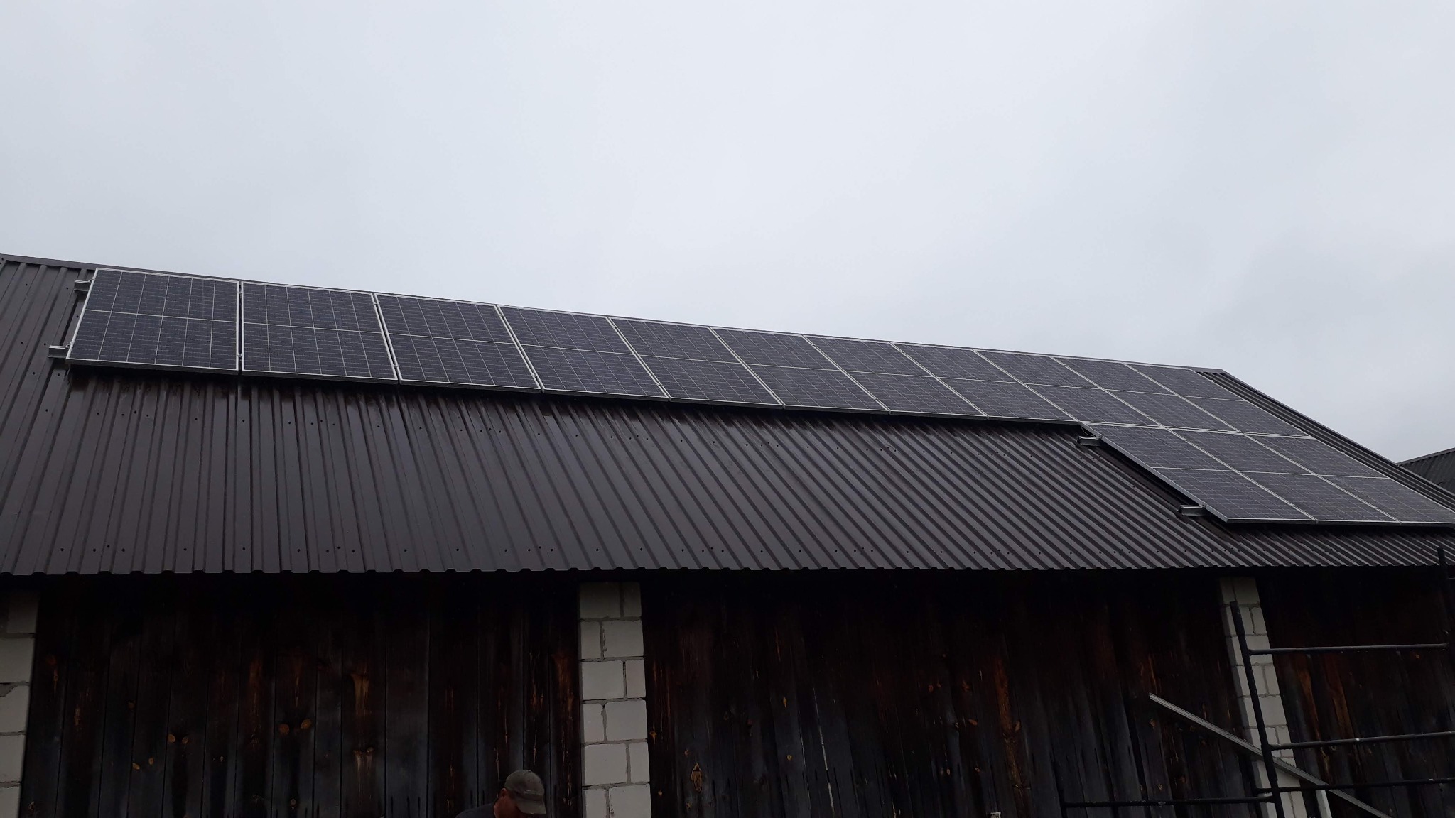 Panele słoneczne zamontowane na ciemnym, metalowym dachu budynku gospodarczego z drewnianymi ścianami i fragmentem rusztowania w dolnym rogu.